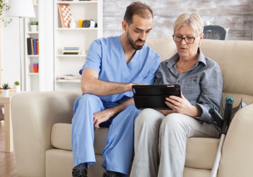 Young doctor in nursing home helping senior woman with crutches to use tablet computer.